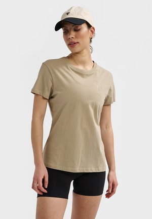 Kvinde iført beige t-shirt med korte ærmer og chevron-logo, sorte cykelshorts og beige og sort kasket mod en ensfarvet baggrund.