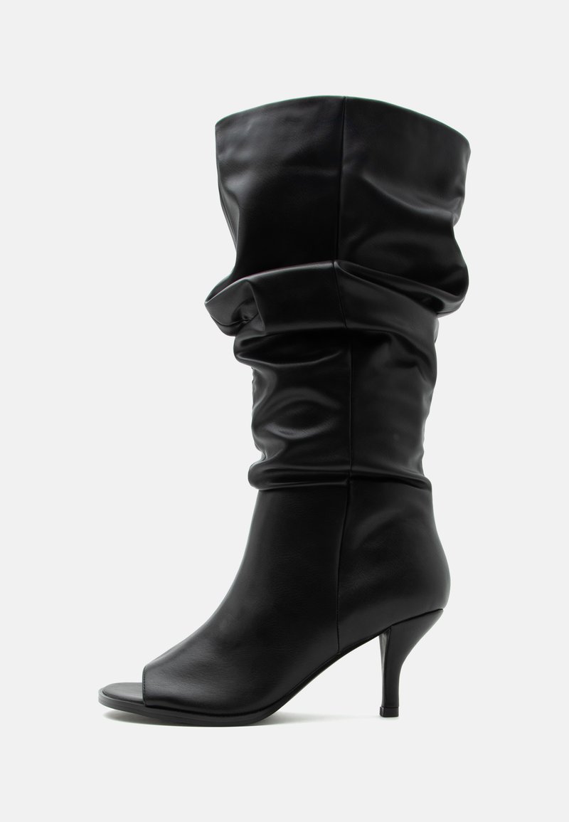 Botte en cuir noire jusqu'au milieu du mollet, ample, avec bout ouvert et talon haut et fin sur fond gris clair uni.