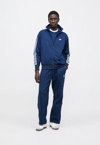 Μπλε ναυτικό φόρμα με ζακέτα με φερμουάρ και λευκές ρίγες στα μανίκια, συνοδευόμενη από παντελόνι στο ίδιο σχέδιο, και τα δύο με λογότυπο. Υφασμένα υλικά με υφή.