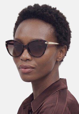 Frau mit kurzen lockigen Haaren, die dunkle verlaufende Cat-Eye-Sonnenbrille und ein braunes Hemd mit Kragen trägt und leicht nach rechts schaut.