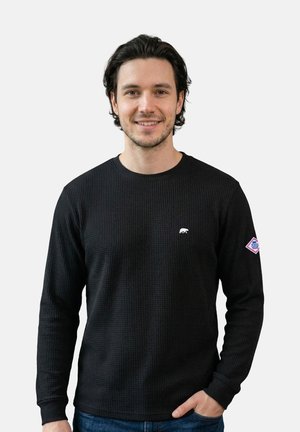 Mann mit dunklem, welligem Haar, der ein schwarzes, strukturiertes Langarmshirt und blaue Jeans trägt und mit einer Hand in der Tasche lächelt.