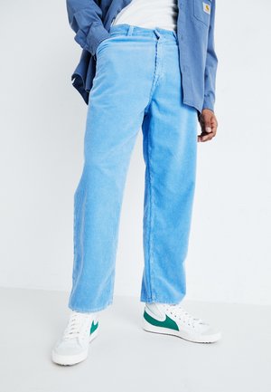 Person iført lyseblå ribbukser, hvide sneakers med grønne accenter, hvid skjorte og blå jakke, stående mod en ensfarvet baggrund.