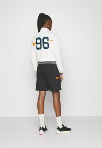 Long-sleeve white top with "96" printed in blue and green, paired with black shorts featuring an orange logo patch and short sleeves. Black sneakers.