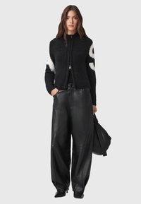 Cardigan nero con zip e motivo bianco sulle maniche, realizzato in materiale morbido; abbinato a pantaloni larghi in pelle nera e una borsa grande.