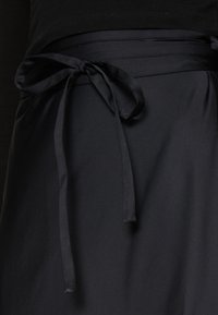 Jupe noire avec une texture lisse, dotée d'une ceinture en satin nouée et d'un design à plusieurs couches. Le tissu semble léger et fluide.