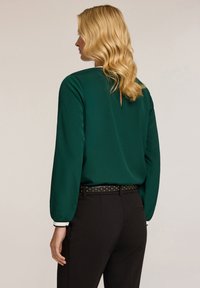 Blusa verde con maniche lunghe, leggermente lucida e dettaglio a occhiello sulla parte posteriore, abbinata a pantaloni neri e una cintura con borchie.