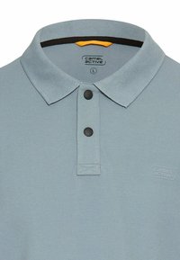 Jasnoniebieska koszulka polo z teksturowanej tkaniny, z zapięciem na guziki z dwiema ciemnymi guzikami i logo po lewej stronie.