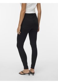 Femme portant un jean skinny noir taille haute et des sandales noires à talons, debout de dos face à l'appareil photo sur un fond blanc.