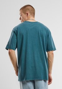 T-shirt unisexe à manches courtes en coton bleu teal délavé. Il présente une coupe décontractée, un col ribba et des détails de couture, avec une étiquette sur une manche.