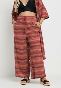 Röda och orange mönstrade vida byxor med dragsko i midjan, med fickor och ett zigzag-mönster, i kombination med en svart top och sandaler.