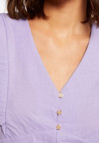 Top in lavanda con scollatura a V e tre bottoni in oro strutturati. Il tessuto sembra leggero, con una texture liscia e un leggero lucido.