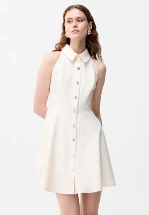 Vrouw met lang golvend haar, gekleed in een mouwloze witte jurk met knopen en kraag, staand met handen achter de rug tegen een effen witte achtergrond.