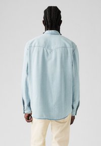 Light blue denim shirt featuring a pointed collar, long sleeves with button cuffs, and a curved hem. Smooth texture with subtle stitching details.