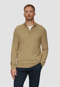 Pull-over zippé beige avec des manches longues, texture douce et poignets côtelés. Porté sur un t-shirt blanc à col rond, associé à un pantalon foncé.