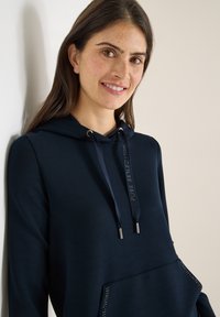 Granatowa bluza z kapturem z przednią kieszenią, wyposażona w sznurek ze metalowymi elementami oraz błyszczący detal na kieszeni. Gładka faktura tkaniny.