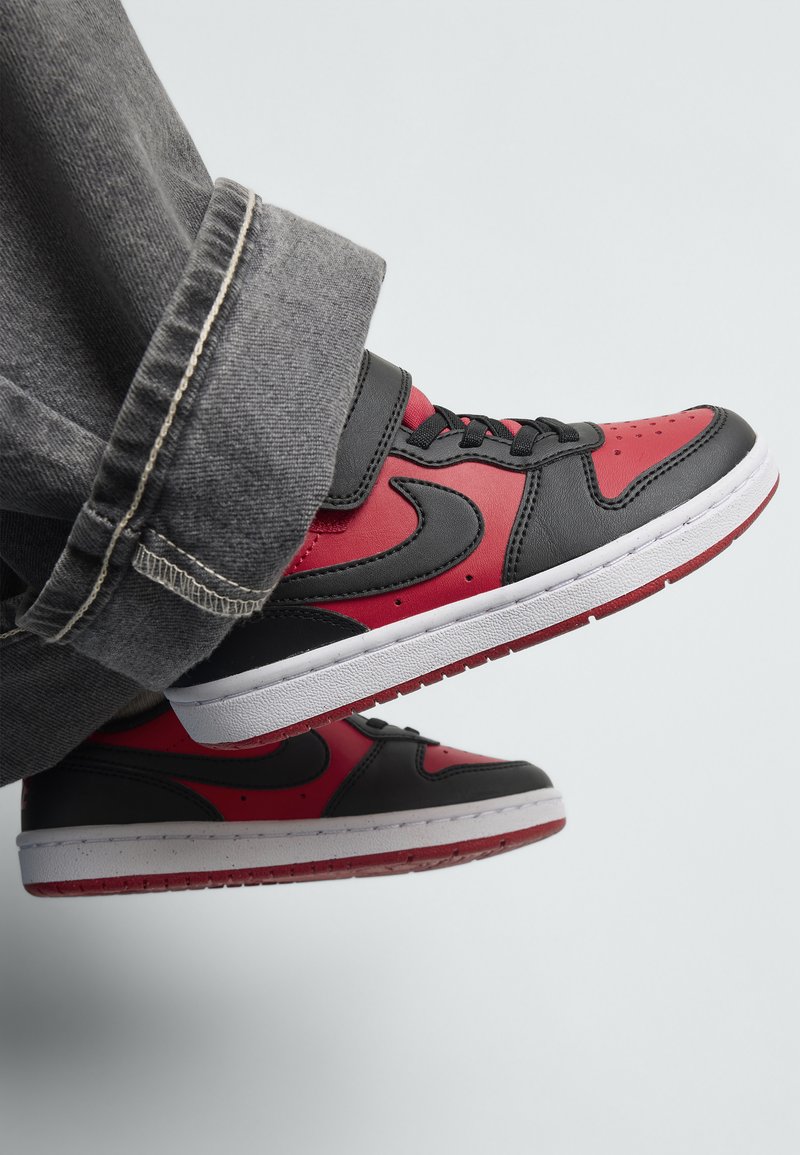 Sorte og røde sneakers med en glat læderoverdel, hvid mellemsål og sorte detaljer. Udstyret med en velcrorem og perforeret tåboks.