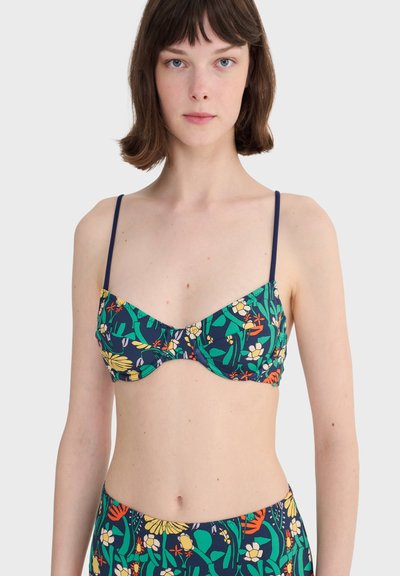 Jeune femme aux cheveux bruns courts portant un haut de bikini bleu marine à fleurs avec de fines bretelles, sur un fond uni.