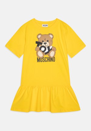 Żółta sukienka z krótkim rękawem i falbaną na dole, z motywem misia trzymającego litery oraz tekstem „MOSCHINO” na froncie.