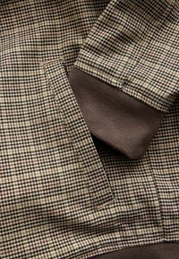 Woven fabric featuring a houndstooth pattern in beige, brown, and black, with a dark brown ribbed cuff detail.