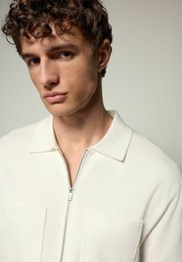 Hvid lynlås sweater med en blød overflade, der har en krave og to frontlommer. Minimalistisk design med en glat overflade.