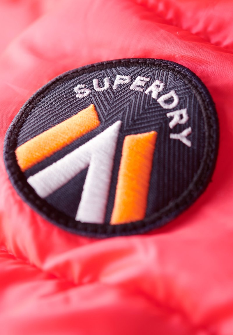Schwarzer, runder Aufnäher mit "SUPERDRY"-Text und einem dreieckigen Design in Weiß und Orange, auf einen roten strukturierten Stoffhintergrund genäht.