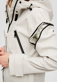 Hellbeige, wasserdichte Jacke mit schwarzen Reißverschlüssen, verstellbare Kapuze und grün gestreiftem Futter, das unter einem hochgerollten Ärmel sichtbar ist.