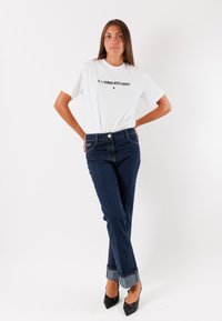 T-shirt bianco con testo nero, abbinato a jeans in denim blu scuro con risvolti. Il modello indossa tacchi neri a punta.