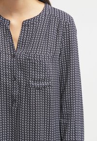 Marinblus med ett geometriskt vitt triangelmönster, långa ärmar, krage som kan vikas ner, knappslå och bröstficka.