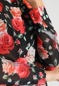 Blouse noire à fleurs avec des roses rouges et roses, en tissu transparent, fermeture à boutons avec des accents métalliques, et manches longues.