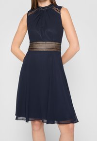 Námořnické modré šaty s plisovaným živůtkem, bez rukávů a ozdobným výřezem v pase. Splývavá sukně s jemnou texturou.