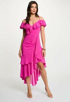 Donna in piedi con un abito fucsia acceso con spalle scoperte, volant e orlo asimmetrico, che indossa braccialetti d'oro e sandali con tacco alto.