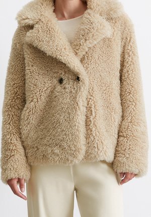 Manteau teddy beige à double boutonnage, douillet, porté sur un haut blanc avec un pantalon crème, doté de larges revers et de manches longues.