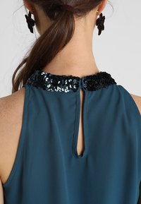 Top senza maniche di colore verde acqua con colletto in paillettes nere, apertura a chiave sul retro e tessuto morbido e leggero.