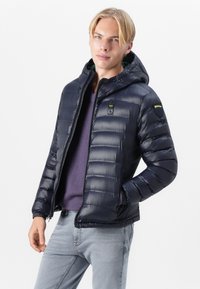 Navy puffer jacket with a hood, quilted design, front zipper, and logo accents. Made of shiny, lightweight material with elastic cuffs.