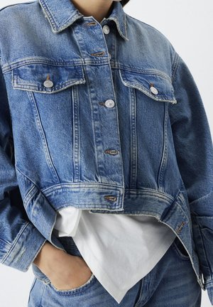 Kobieta w niebieskiej kurtce dżinsowej z metalowymi guzikami na białej koszuli, z ręką swobodnie w kieszeni dżinsów na jednolitym tle.