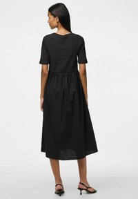 Crna haljina s tijesnim korzetom i kratkim rukavima, koja prelazi u labavu, naboranu suknju. Mekani, prozračni materijal s suptilnom teksturom.