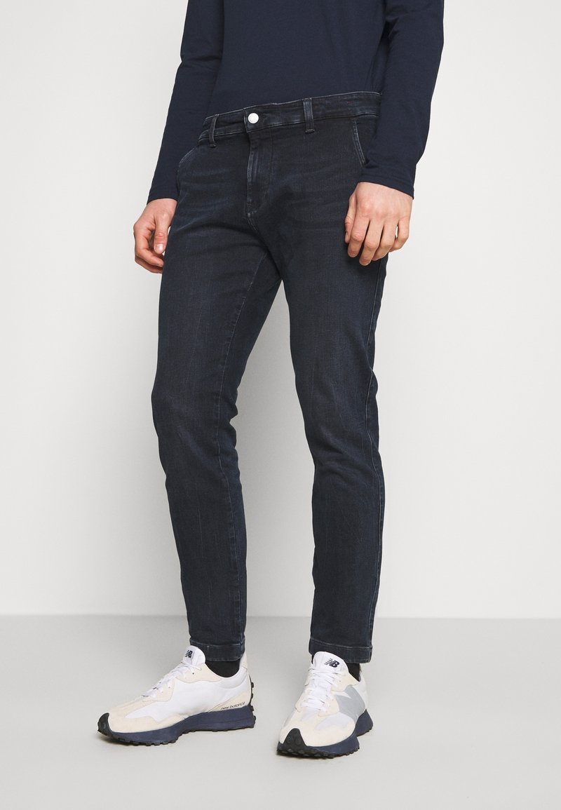 Mörkblå denimjeans med slim fit, som har fem fickor och knäppning fram, vilka bärs med vita och grå sneakers.