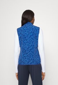 Kvinna med långt mörkt hår som bär en blåmönstrad ärmlös väst över en vit långärmad skjorta och mörka byxor, sedd bakifrån.