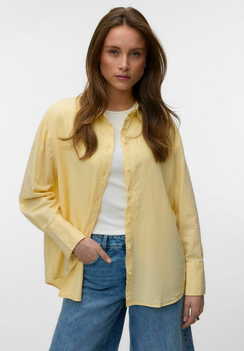 Jeune femme aux longs cheveux bruns porte une chemise jaune ample à boutons sur un haut blanc et un jean bleu, debout avec une main dans la poche.