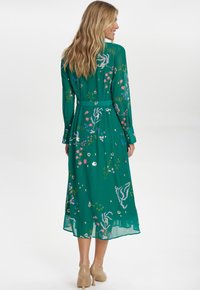 Hosszú zöld ruha virágmintával, különböző színekben, hosszú ujjakkal, övvel a deréknál és áramló, félig átlátszó anyaggal.