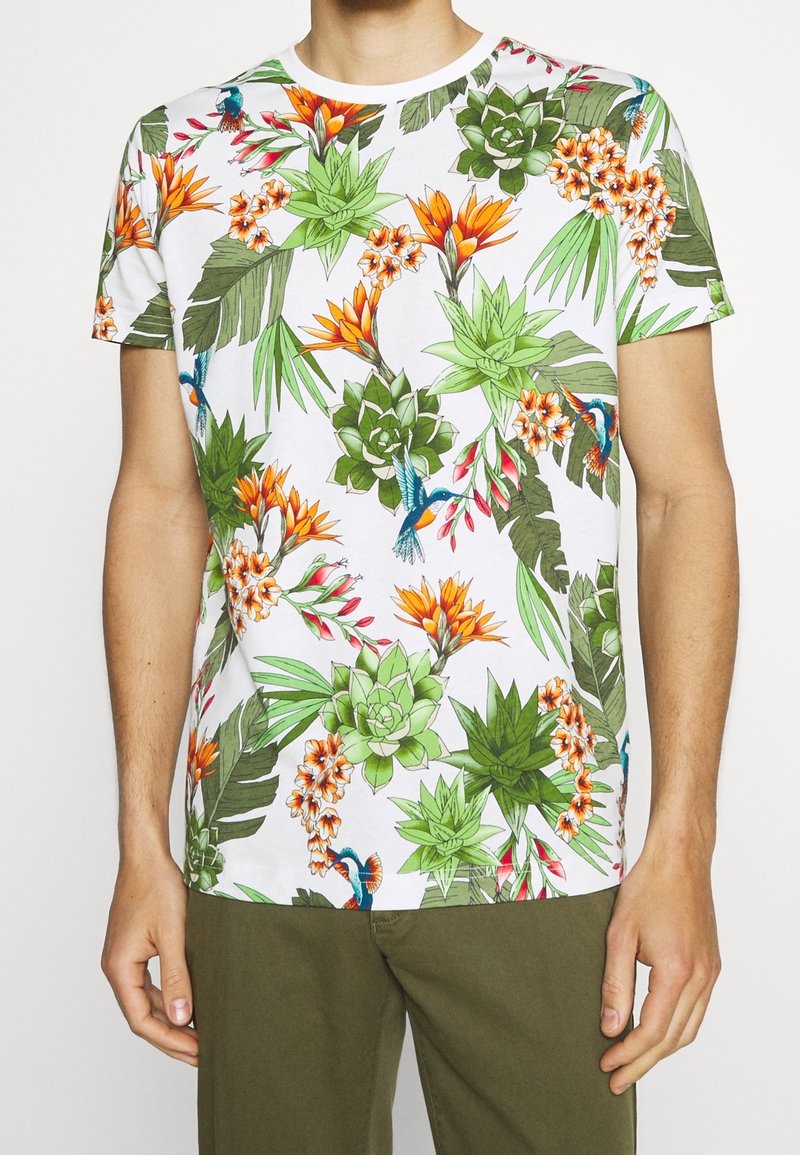 Camiseta de manga corta y cuello redondo con un vibrante estampado tropical de flores y follaje en verde, naranja y azul sobre un fondo blanco.