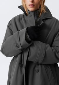 Cappotto grigio di grandi dimensioni con colletto alto, grandi bottoni e maniche larghe. Indossato sopra un maglione scuro e testurizzato e guanti neri.
