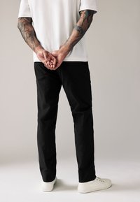 Pantalones negros de corte recto, hechos de una tela suave. Se llevan con una camisa blanca y zapatillas blancas. Tatuajes visibles en los brazos.