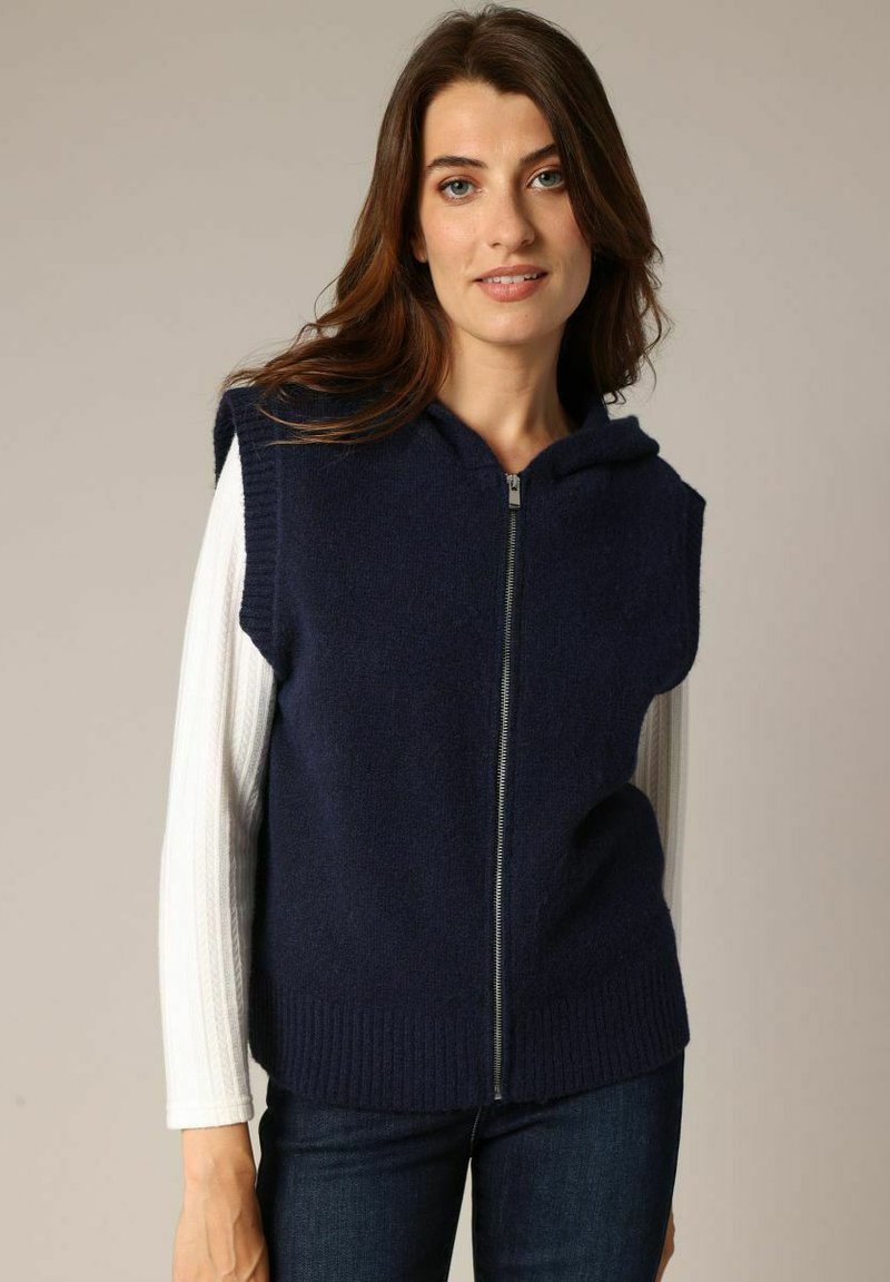 Gilet tricoté bleu marine avec un devant zippé et une capuche, présentant un ourlet côtelé et des manches courtes, porté par-dessus un haut à manches longues blanc.