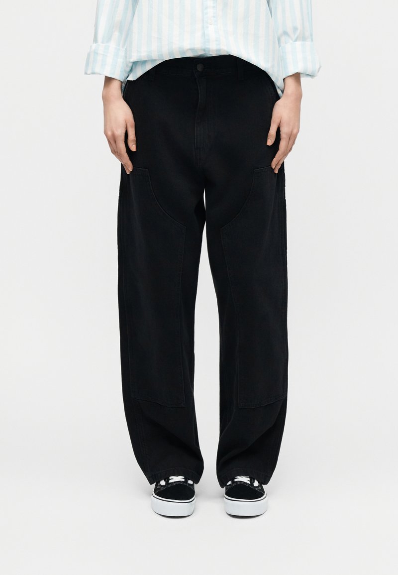 Personne portant un pantalon noir ample avec de grandes poches frontales, des baskets blanches et noires, et une chemise à manches longues bleu clair à rayures.