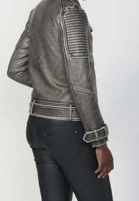 Giacca in finta pelle grigia con dettagli a coste sulle spalle, strisce verticali sulle maniche e vita con cintura. Indossata con pantaloni in pelle neri aderenti.