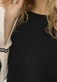 Pull en maille noire avec un motif texturé, partiellement recouvert d'un cardigan crème avec des rayures noires et des designs géométriques.