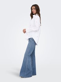 Nuori nainen, jolla on pitkät ruskeat hiukset, pukeutuneena valkoiseen kukkakuosiseen paitaan ja korkeavyötäröisiin leveälahkeisiin sinisiin farkkuihin, seisoo profiilissa valkoista taustaa vasten.