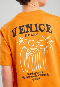 T-shirt in cotone arancione con grafiche nere sul retro, tra cui "VENICE surf shop", tavole da surf, palme e un sole.