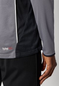 Grå og svart treningsskjorte med mesh-texturer; har en total90-logo og hvite sømmer langs kantene. Nærbilde av ermet og siden.